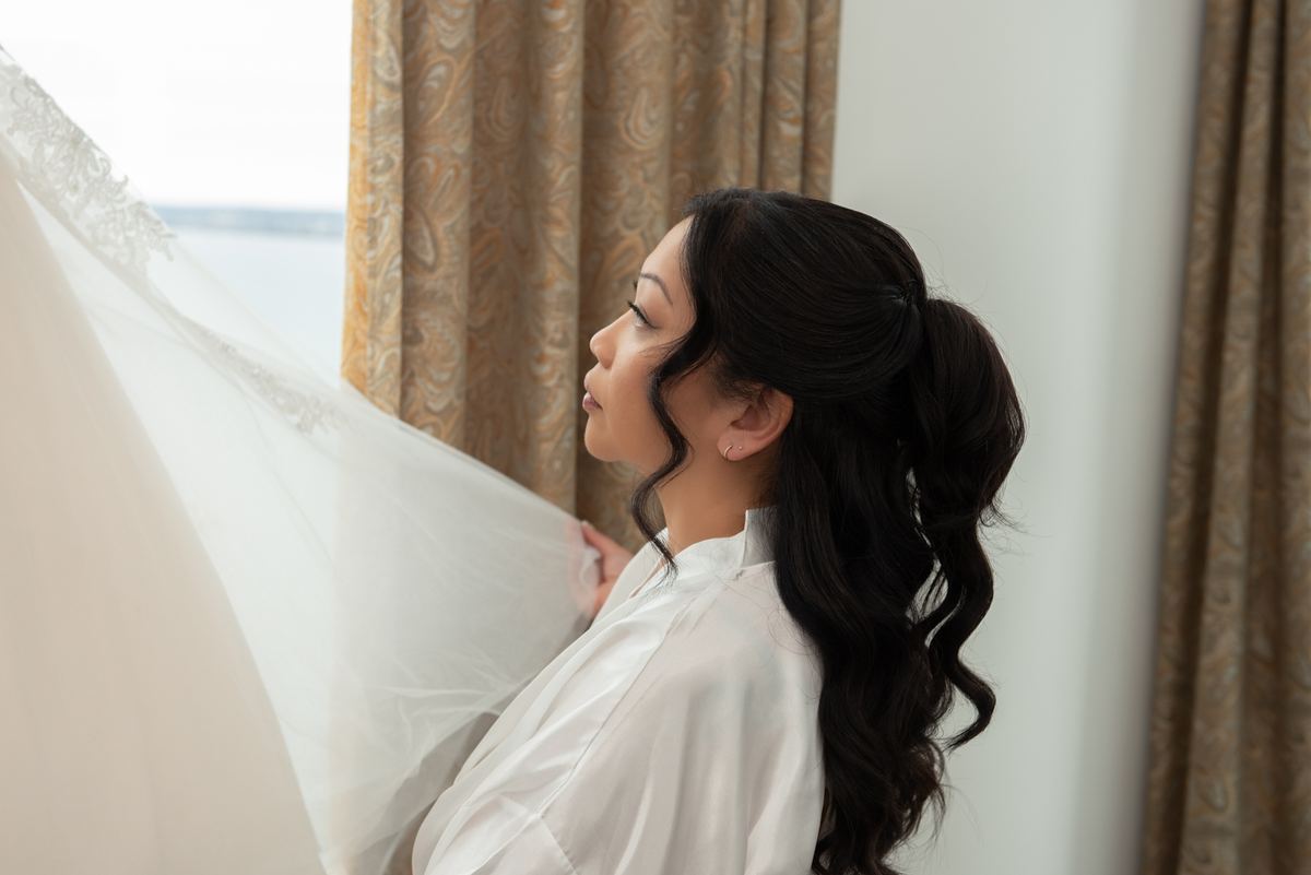 A bride in a white robe stands by a window, holding her veil as she gazes at a hazy waterfront bathed in soft daylight.