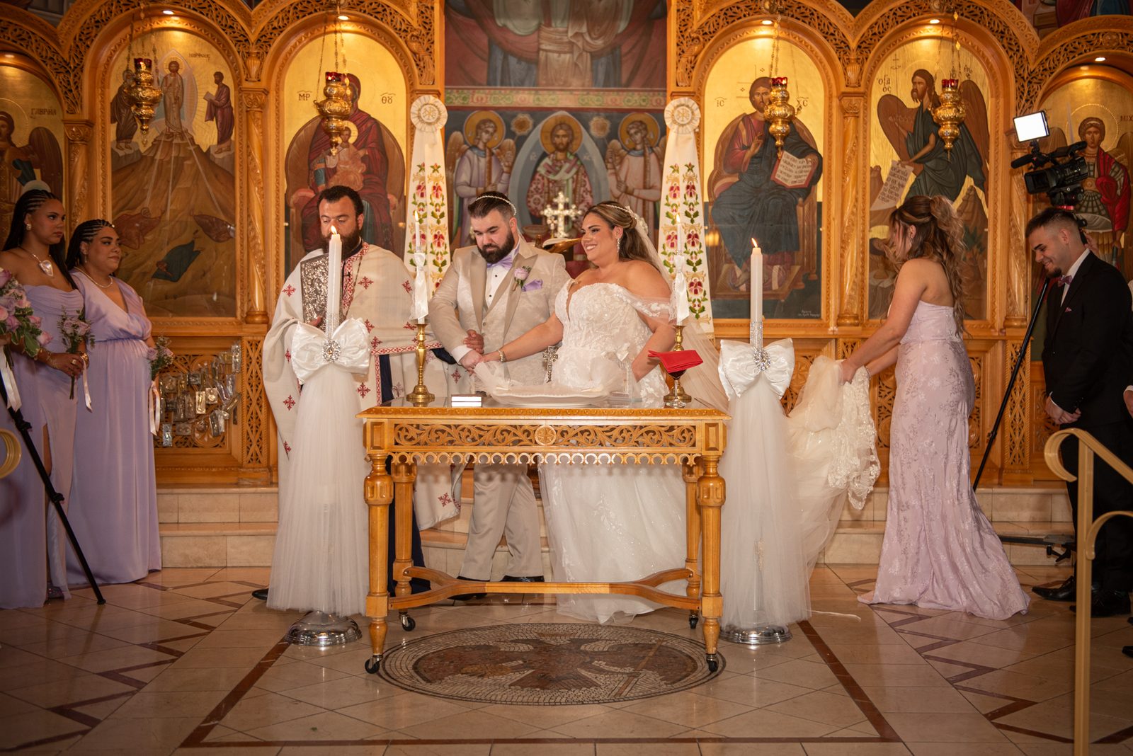 Bride and groom light candles at an ornate altar in an Eastern Orthodox church surrounded by clergy and bridesmaids.