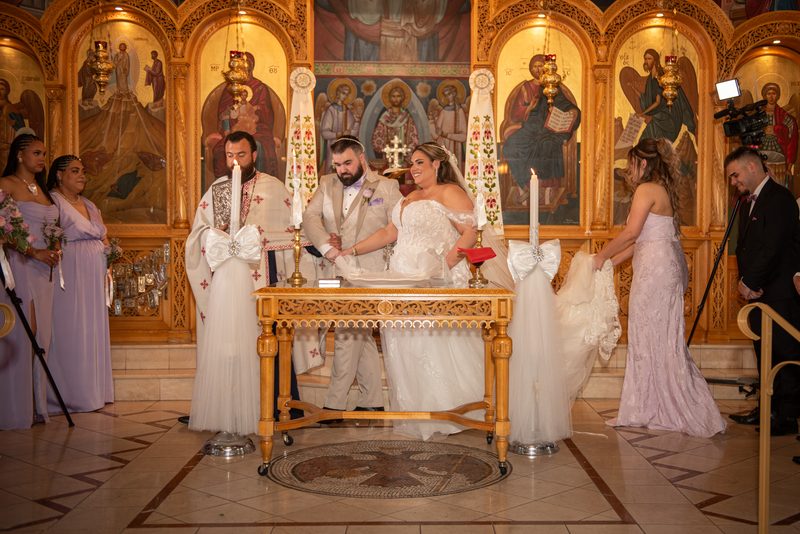 Bride and groom light candles at an ornate altar in an Eastern Orthodox church surrounded by clergy and bridesmaids.