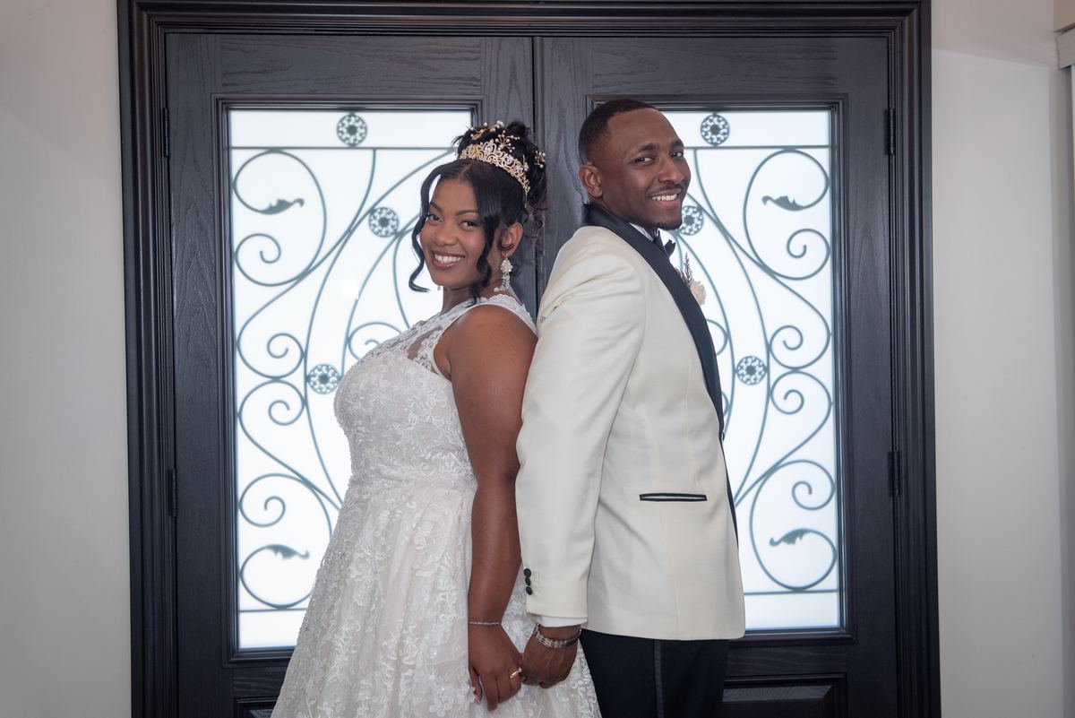 A smiling bride and groom stand back-to-back, hands clasped, in front of an ornate glass door, illuminated by soft light.