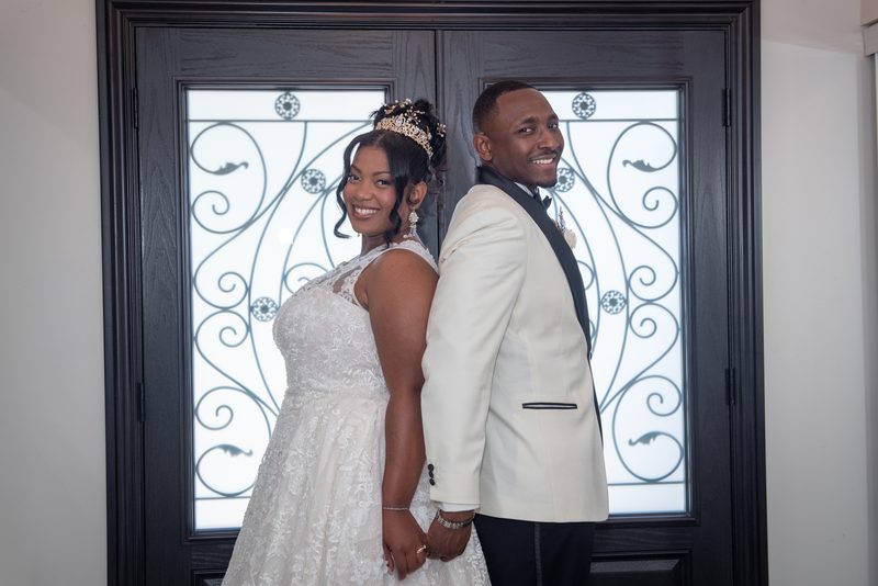 A smiling bride and groom stand back-to-back, hands clasped, in front of an ornate glass door, illuminated by soft light.
