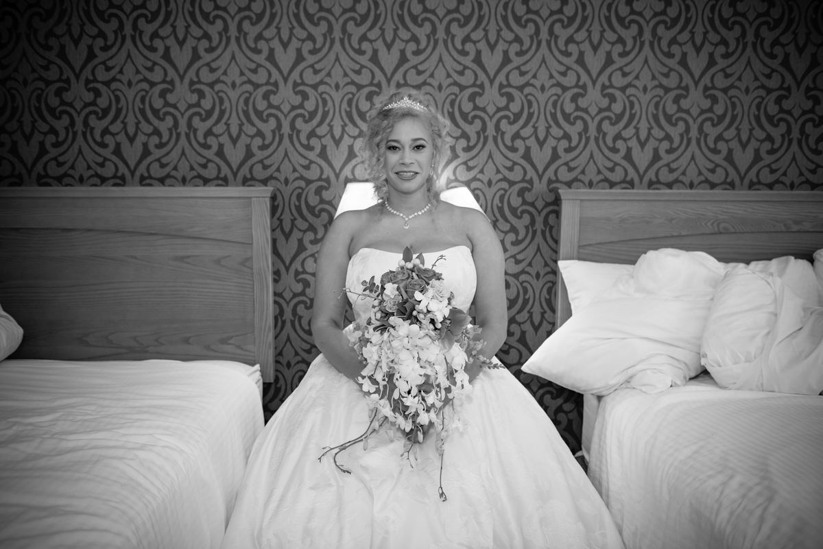 A smiling bride in a wedding gown holds flowers between two beds in a hotel room with ornate wallpaper.