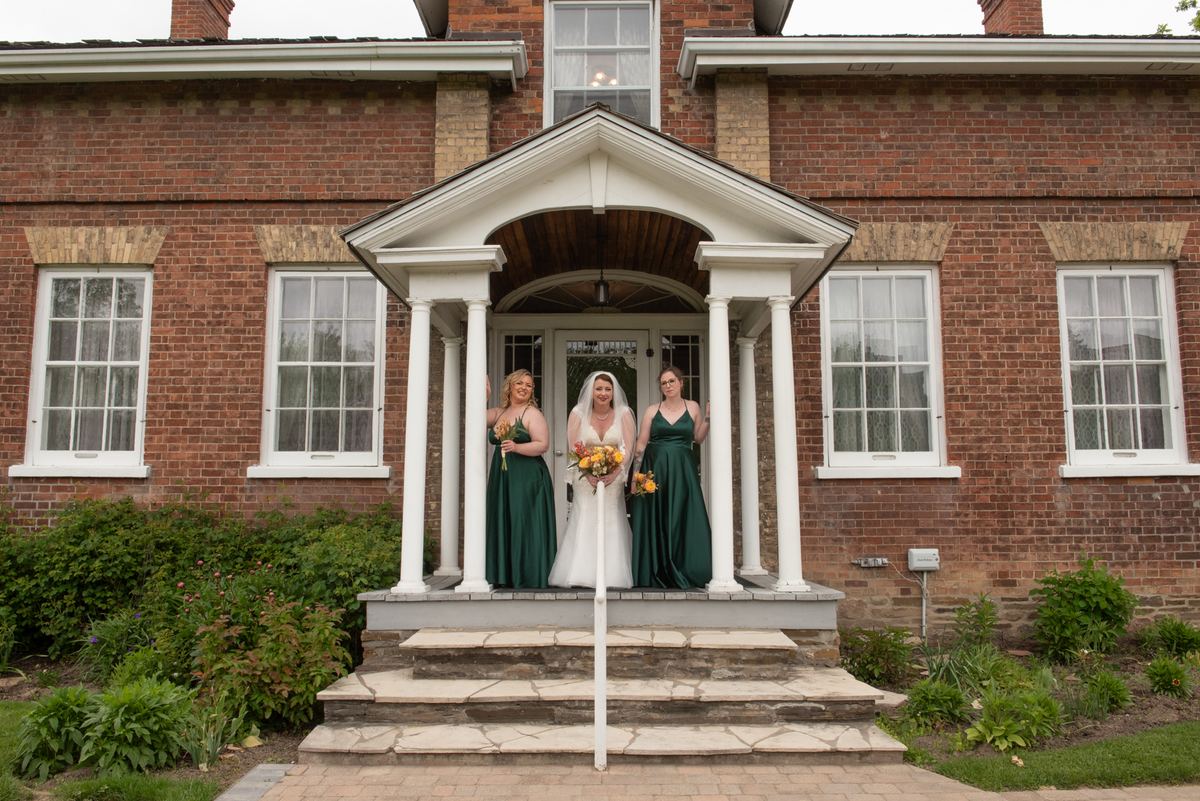 A bride in white stands on stone steps with two bridesmaids in emerald green dresses holding bouquets.