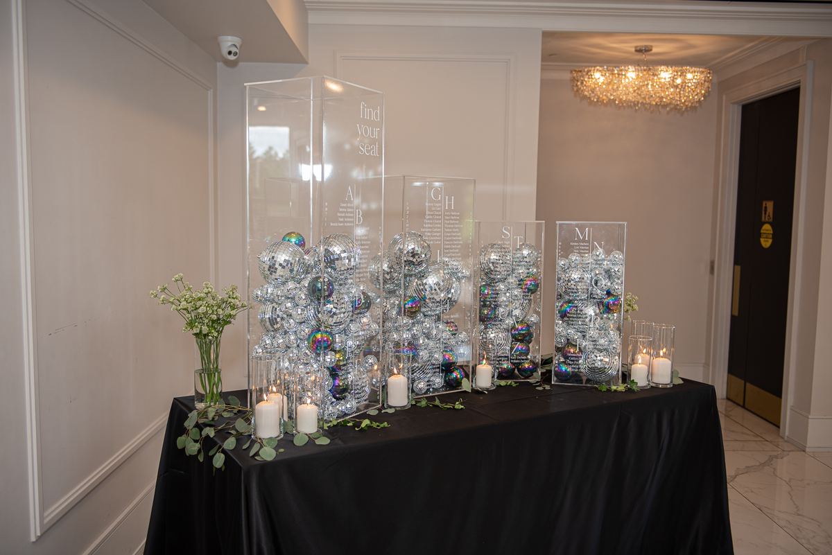 A black-draped table with acrylic stands labeled A–B, G–H filled with disco balls and surrounded by candles and flowers.