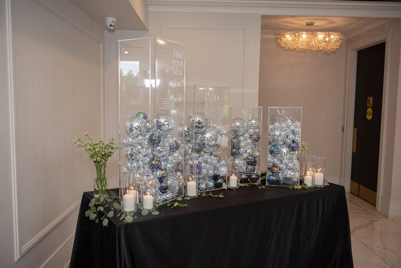 A black-draped table with acrylic stands labeled A–B, G–H filled with disco balls and surrounded by candles and flowers.