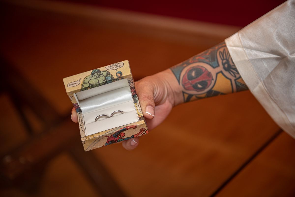 A person with a comic book tattooed forearm opens a ring box revealing two wedding bands, centered in a softly lit frame.