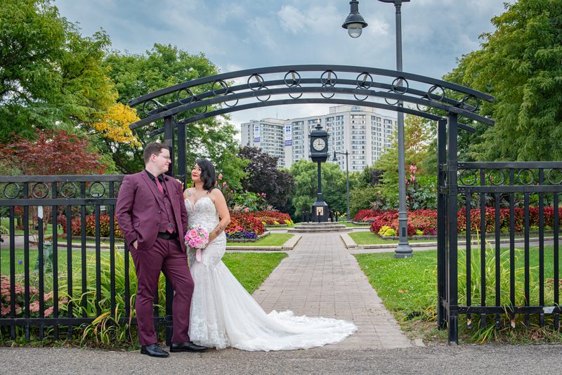 A bride and groom pose under an ornate iron arch in a park with manicured gardens and a clock tower visible behind them.