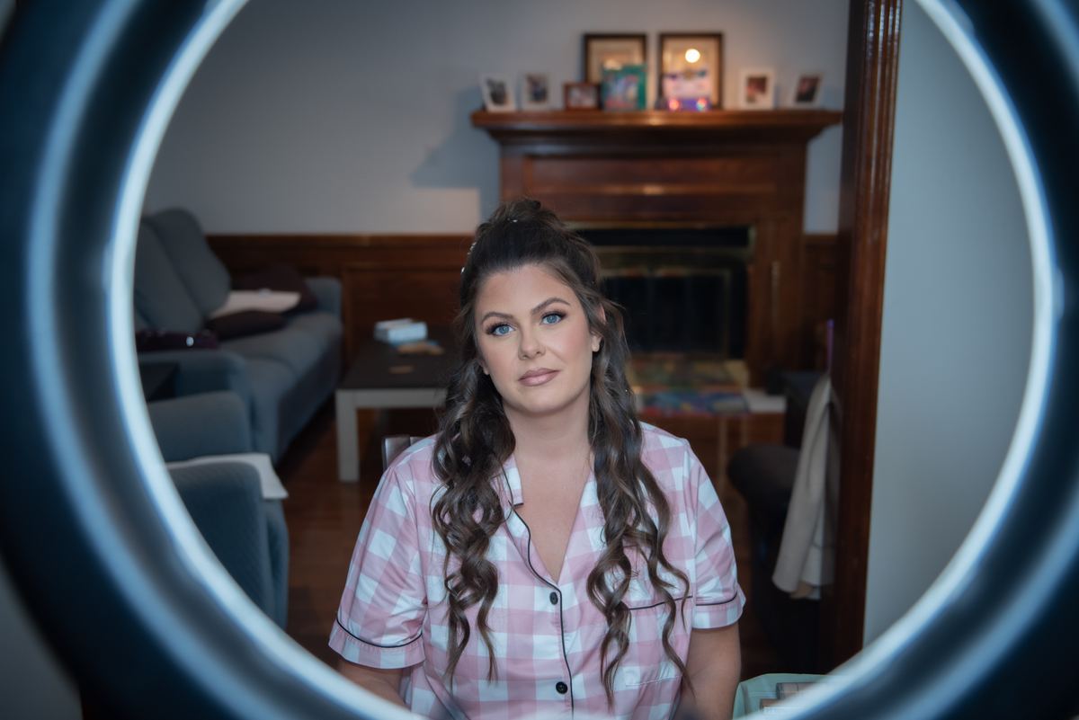 A young woman in pink and white pajamas sits calmly in front of a ring light, gazing directly at the camera.