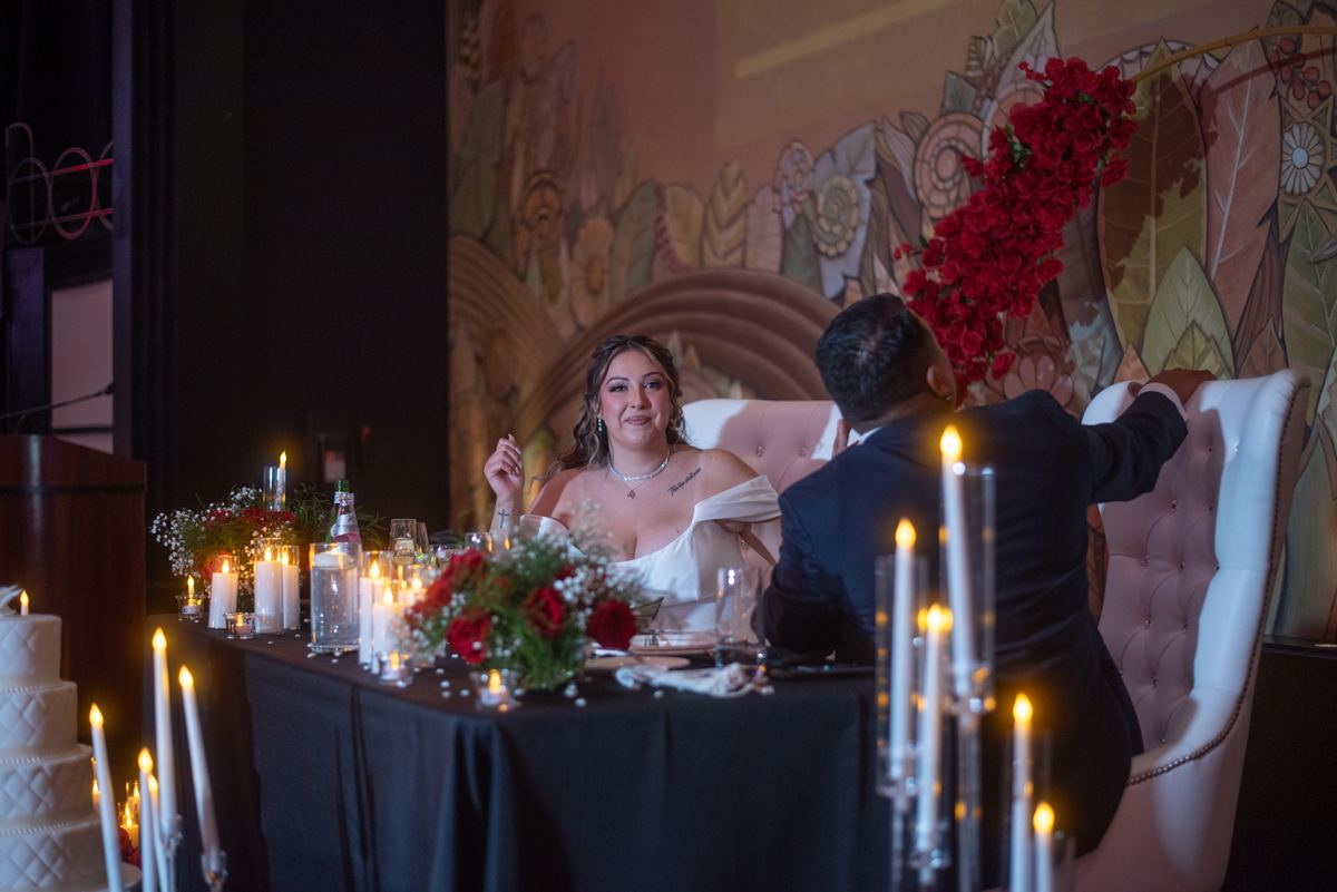 Bride smiles at groom across candlelit table in romantic setting with floral arrangements and mural.