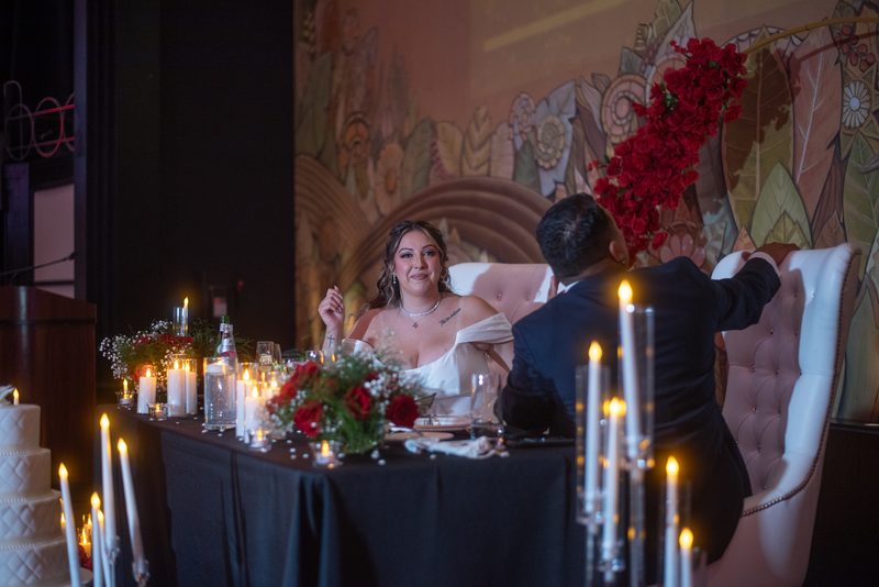 Bride smiles at groom across candlelit table in romantic setting with floral arrangements and mural.