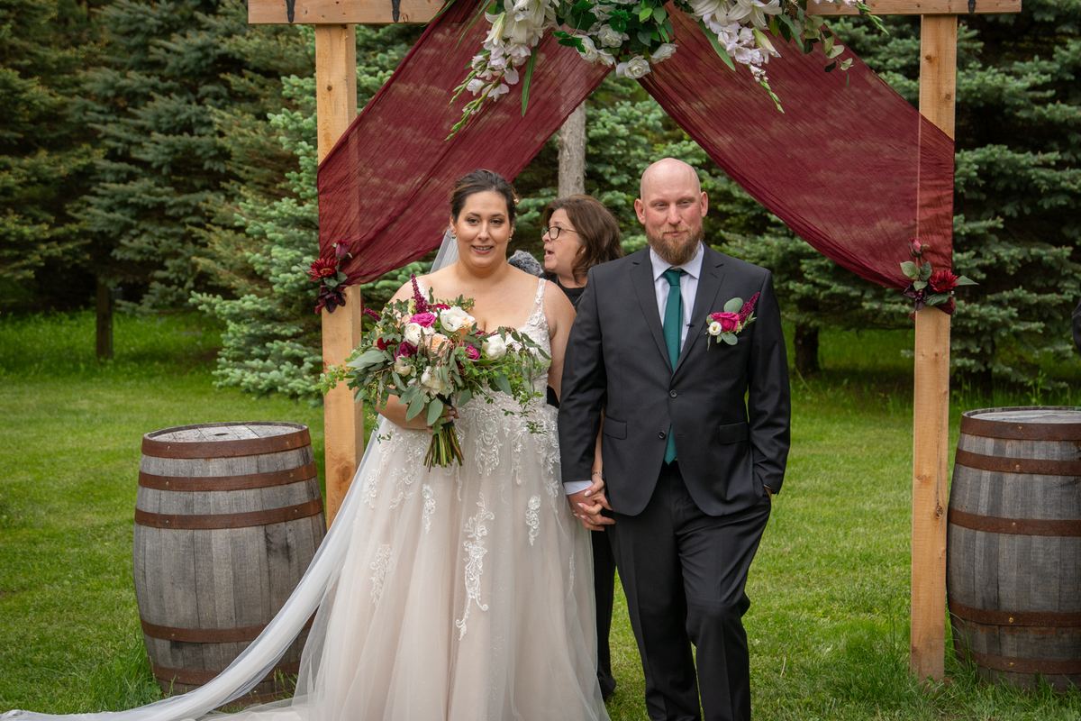 Bride and groom stand under a wooden arch with burgundy drapes and flowers, on a grassy lawn with evergreen trees.