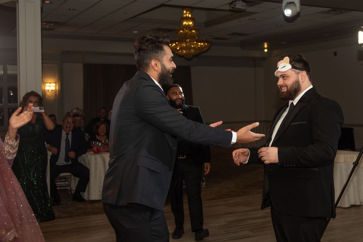 Two men in suits engage in a playful handshake, one wearing a headband, amid laughing guests and warm lighting.