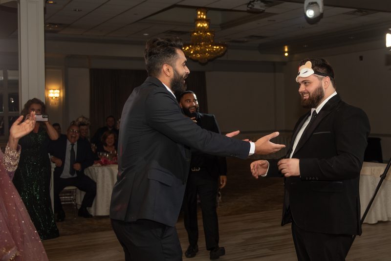 Two men in suits engage in a playful handshake, one wearing a headband, amid laughing guests and warm lighting.