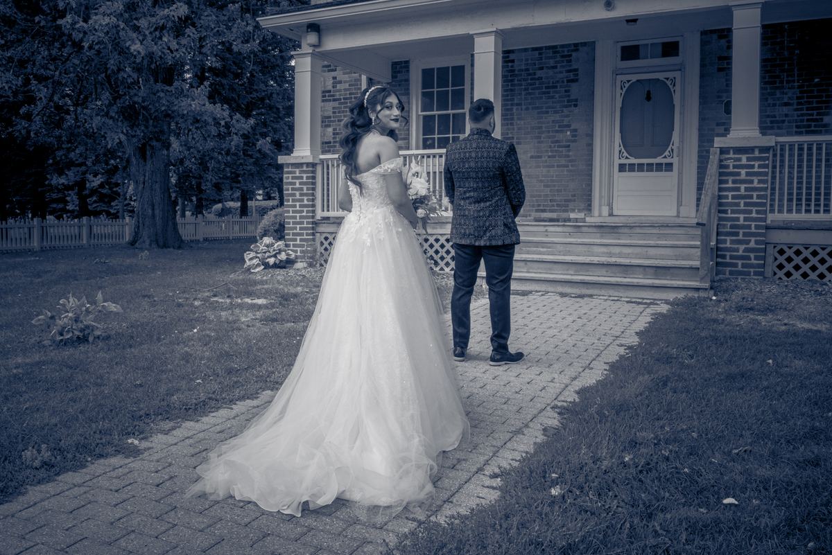 A bride in white and groom in a patterned jacket stand beside a house's front door, awaiting entry on a cool, overcast day.