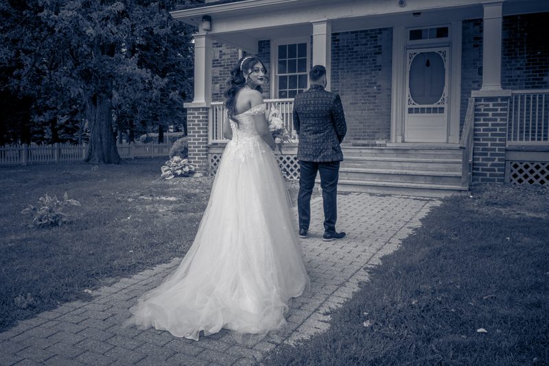 A bride in white and groom in a patterned jacket stand beside a house's front door, awaiting entry on a cool, overcast day.
