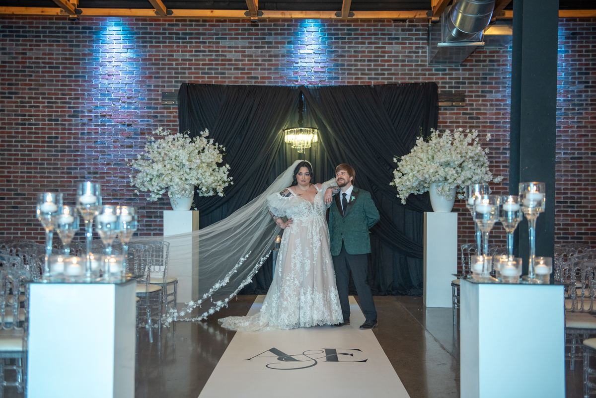 A bride in a lace gown and veil stands with her groom in a green suit at the end of an aisle, surrounded by uplighting.