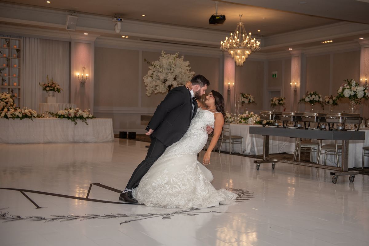 A bride and groom dance closely in an elegantly lit ballroom, surrounded by floral arrangements and set tables.
