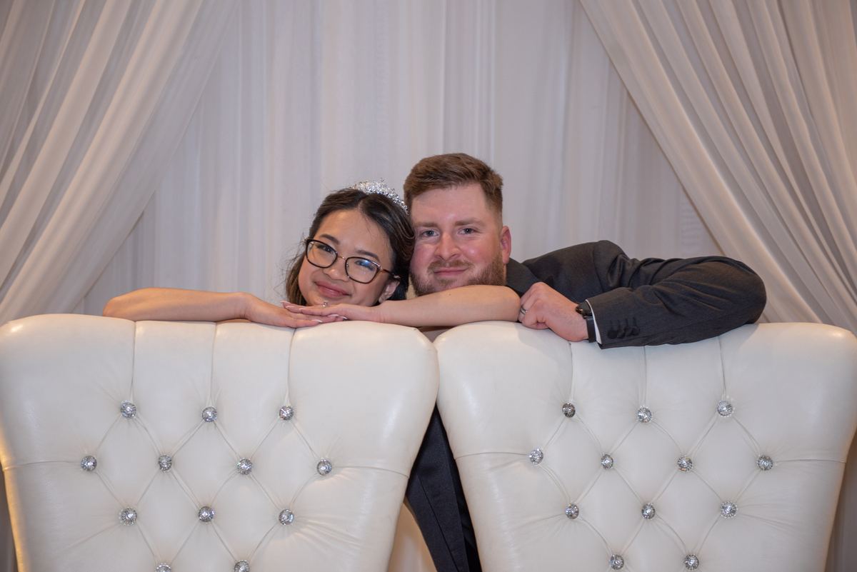 A smiling couple leans on a white tufted bench with crystal buttons, framed by draped white curtains.