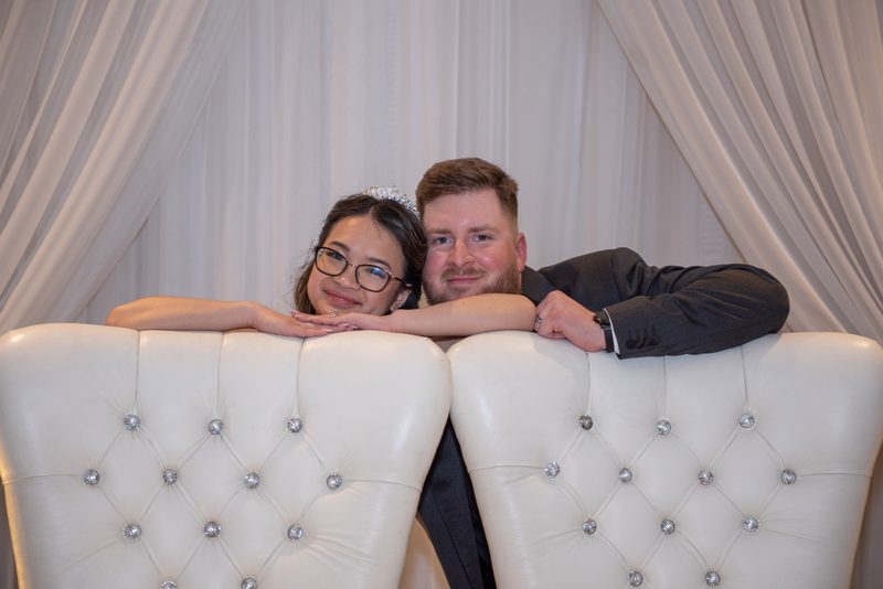 A smiling couple leans on a white tufted bench with crystal buttons, framed by draped white curtains.