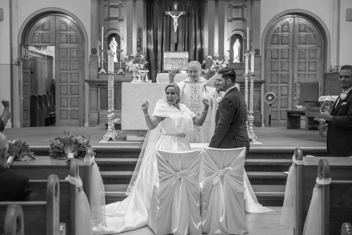 Bride raises her fist joyfully as groom turns to face her, both smiling at church altar surrounded by guests.