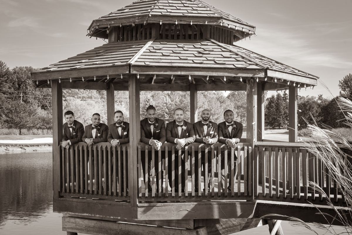 Seven groomsmen in tuxedos smile on a wooden gazebo overlooking water, framed by trees and reeds in sepia tone.