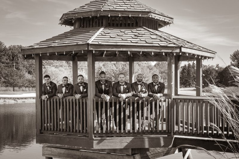 Seven groomsmen in tuxedos smile on a wooden gazebo overlooking water, framed by trees and reeds in sepia tone.