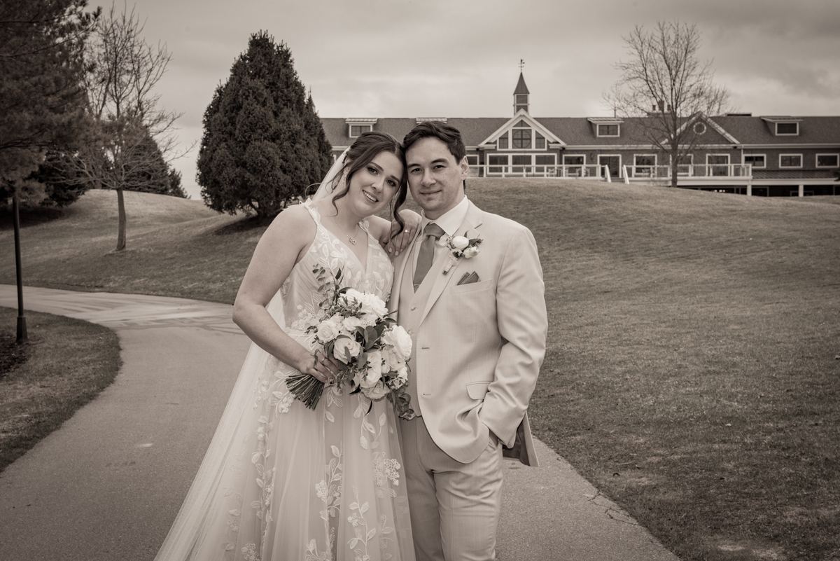 Newlyweds smile for a sepia-toned portrait on a paved path with a stately building in the background.