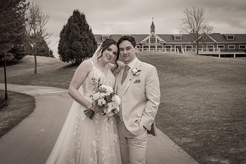 Newlyweds smile for a sepia-toned portrait on a paved path with a stately building in the background.