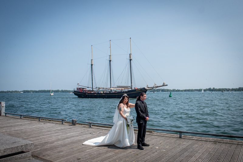 A bride and groom stand on a wooden pier by water, with a sailing ship in the background during their photoshoot.