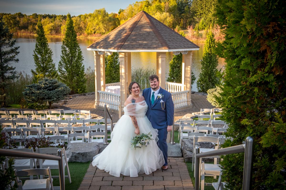 A smiling newlywed couple stands on a brick path near a lakeside gazebo, surrounded by white chairs and autumn trees.