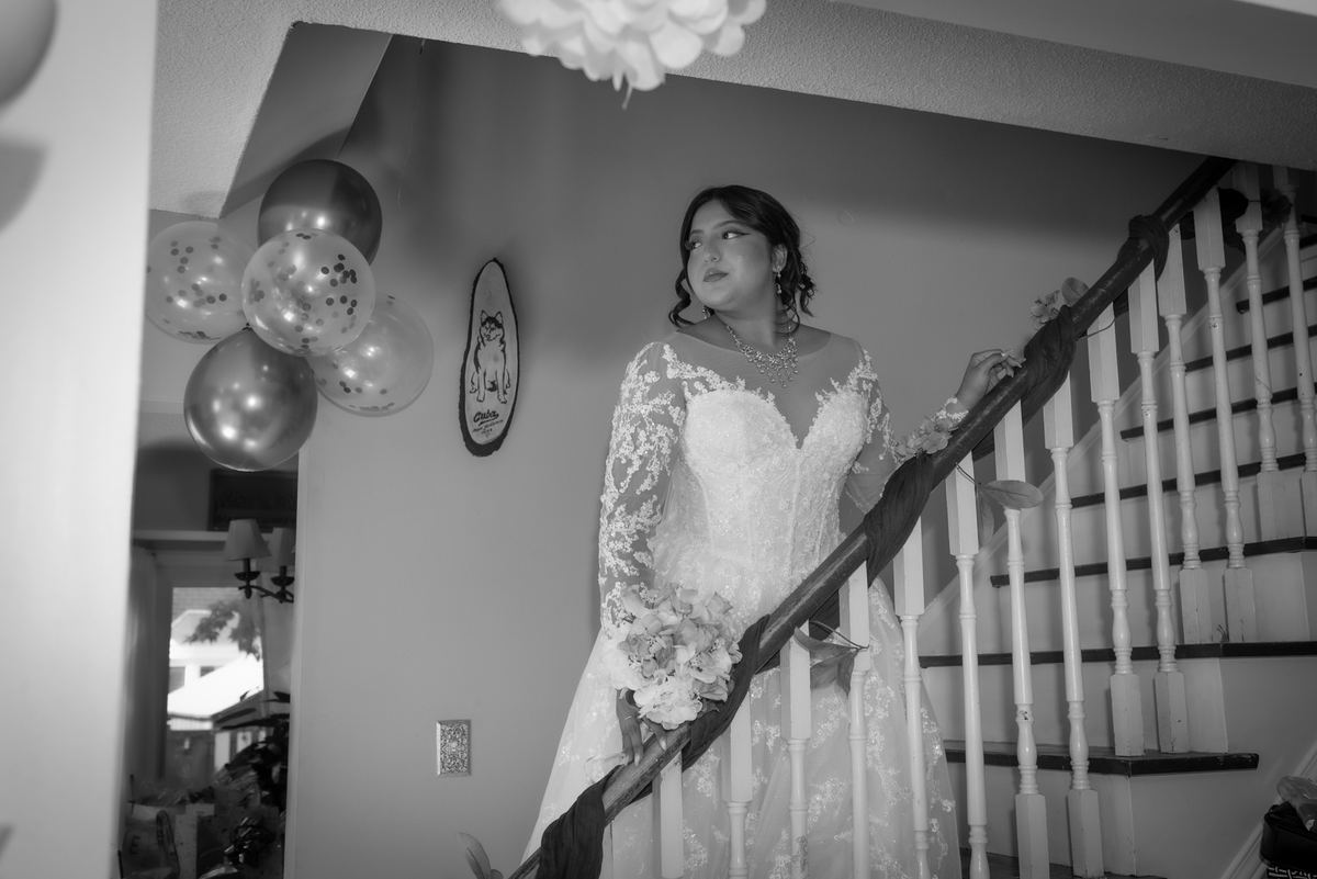 A bride in an ornate lace gown stands contemplatively on a staircase, surrounded by festive balloons and a decorative plaque.