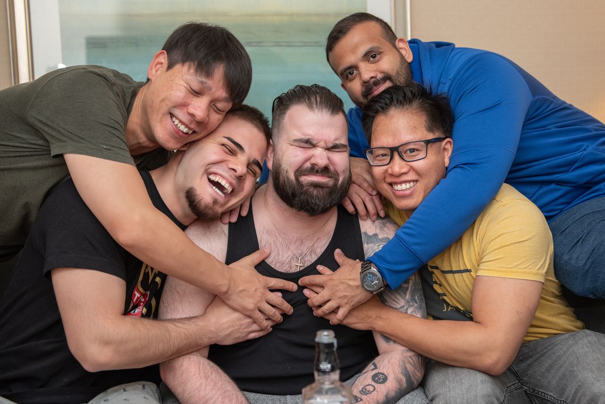 Five men laugh joyfully together, one man's face contorted with laughter in an intimate indoor setting.