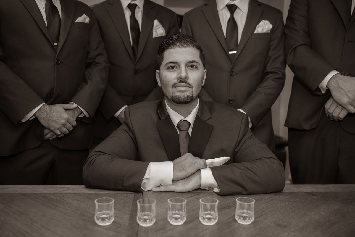 A groom sits at a wooden table with four shot glasses, surrounded by his suited groomsmen, capturing a moment of unity.