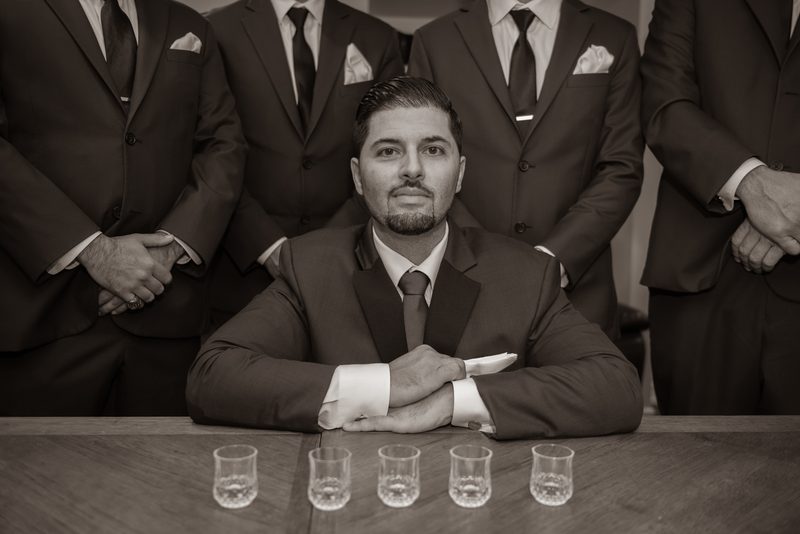 A groom sits at a wooden table with four shot glasses, surrounded by his suited groomsmen, capturing a moment of unity.