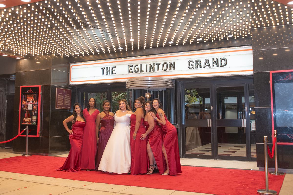 A bride in white with red-dressed bridesmaids pose joyfully outside under string lights and the theater's marquee.