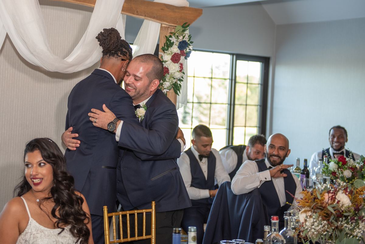 Two grooms embrace warmly at their reception, surrounded by smiling guests in formal attire and elegant table settings.