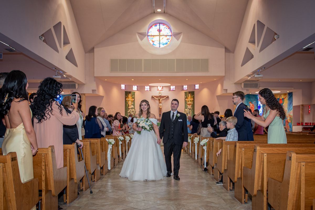 Bride and groom walk down the aisle of a modern church, illuminated by warm light from a rose window.