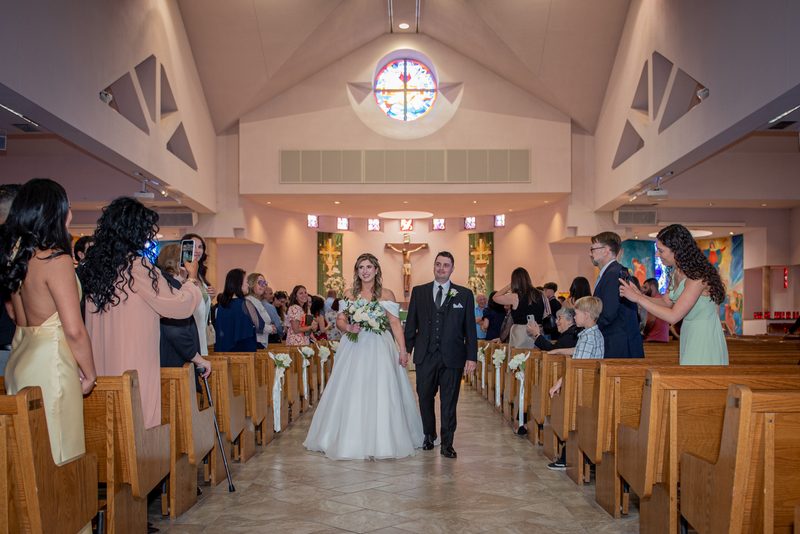 Bride and groom walk down the aisle of a modern church, illuminated by warm light from a rose window.