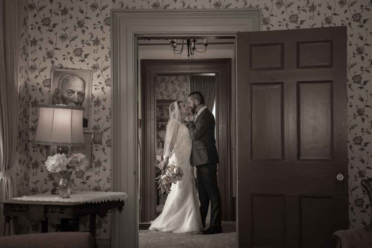 A bride and groom kiss in a vintage-style room with an open doorway, the soft light highlighting their intimate moment.