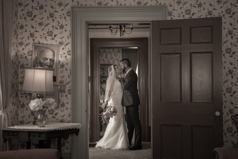 A bride and groom kiss in a vintage-style room with an open doorway, the soft light highlighting their intimate moment.