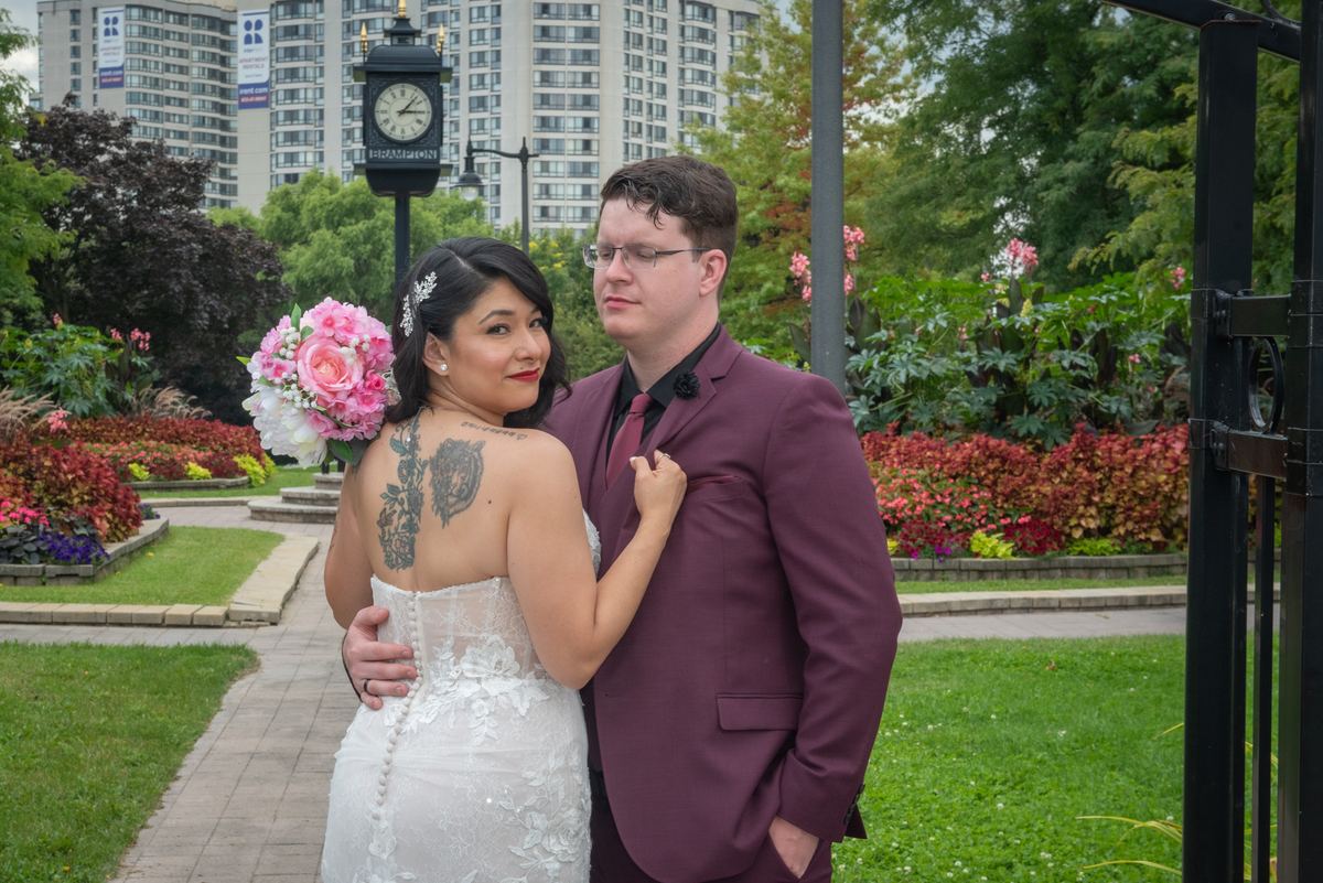 A bride in a lace gown and groom in a burgundy suit pose in a park with gardens and a clock tower behind them.