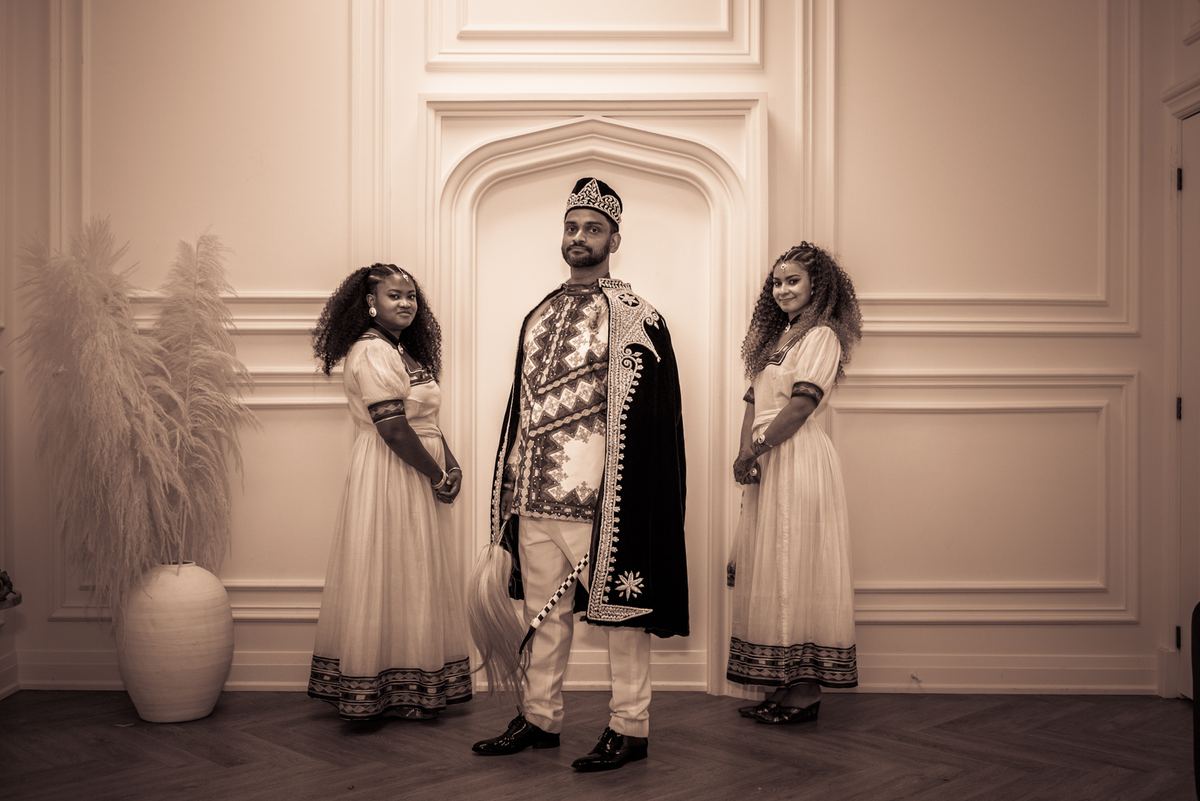 A man in a ceremonial crown and robe stands between two women in matching traditional dresses in an elegant paneled room.
