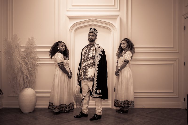 A man in a ceremonial crown and robe stands between two women in matching traditional dresses in an elegant paneled room.