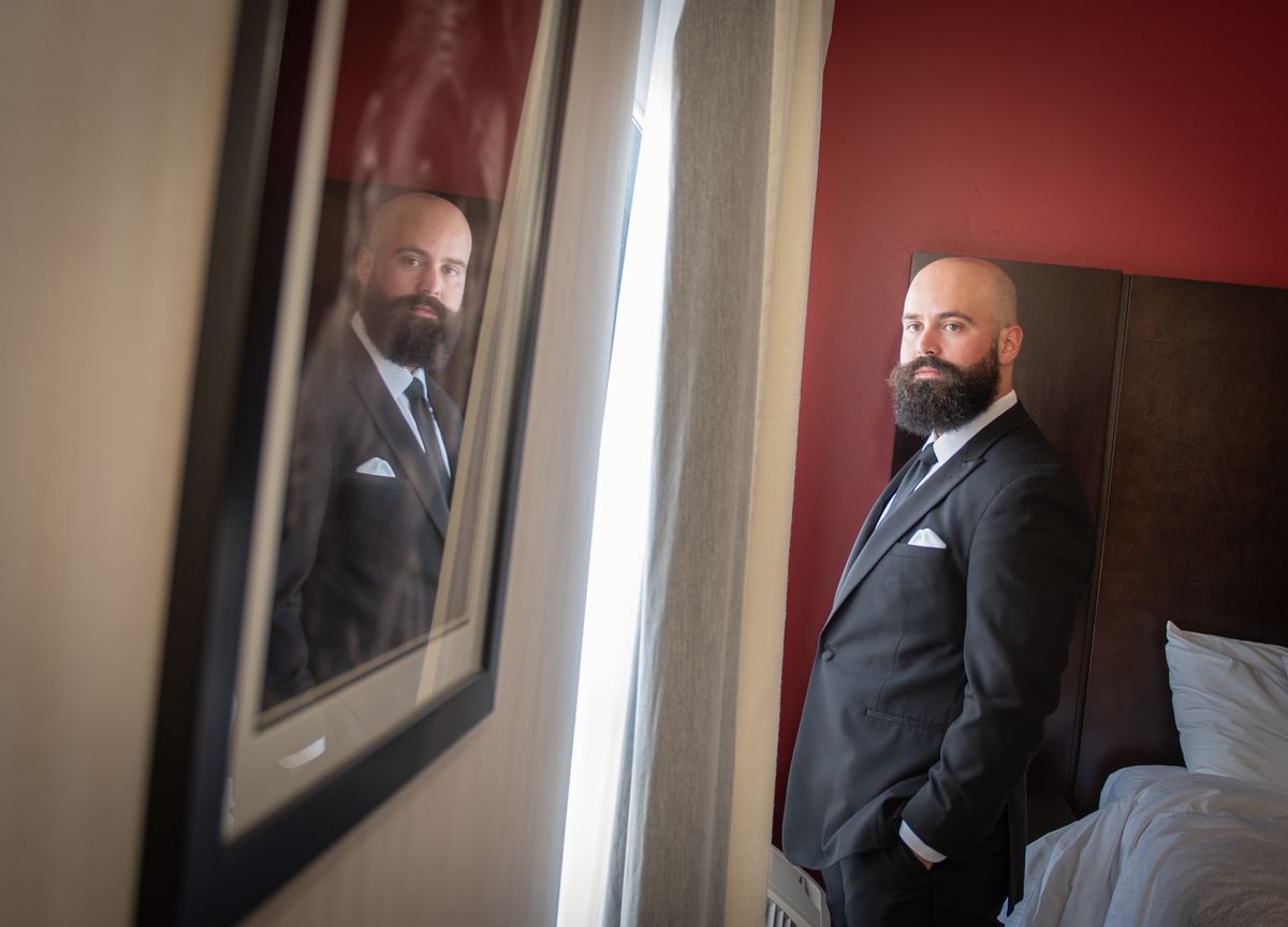 A groom in a suit stands by a window, thoughtfully looking at his portrait on the wall, anticipation.