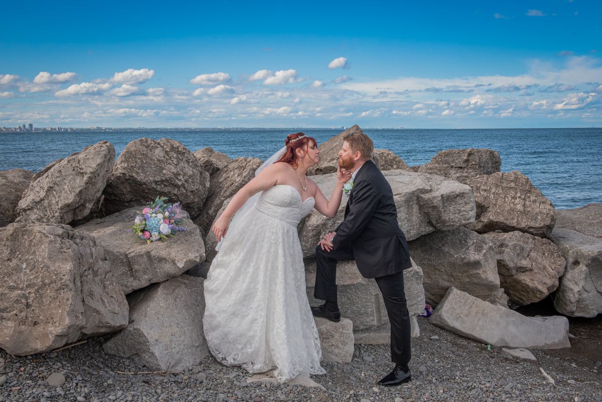 A bride and groom share an affectionate moment by the water's edge, with a city skyline in the background.