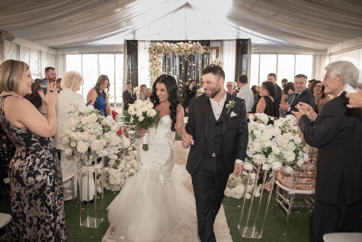 Bride and groom walk down an aisle lined with white flowers, smiling as guests clap under a bright tent.