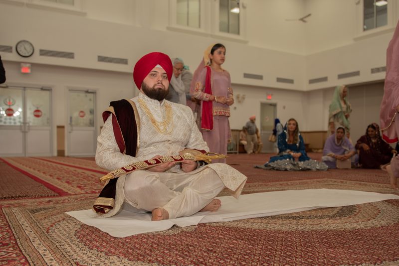 A Sikh groom in white and maroon attire with a red turban sits cross-legged on a white cloth inside a gurdwara.