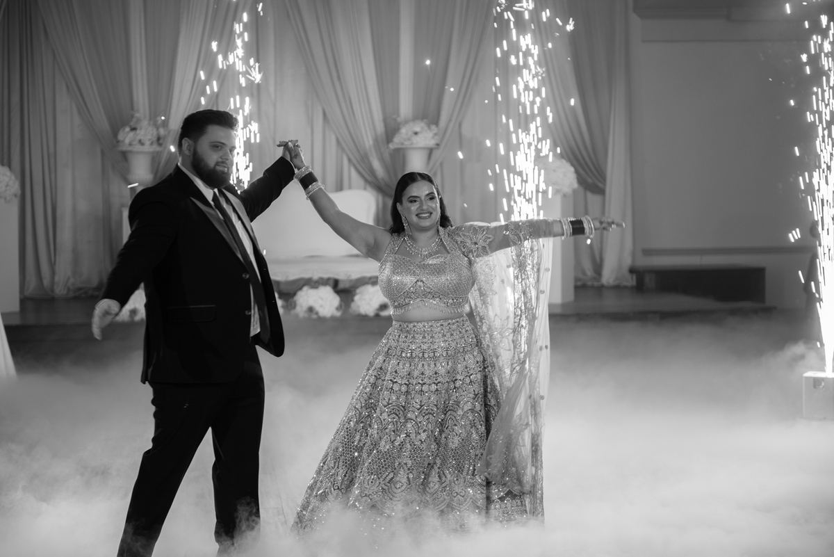 A newlywed couple dances joyfully in an indoor space filled with smoke and illuminated by cascading sparklers.