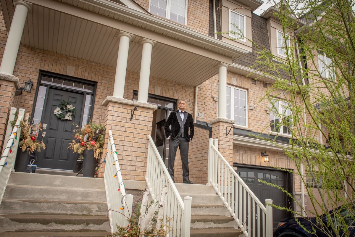 A man in a black tuxedo stands on the steps of a modern brick townhouse, hands in pockets, gazing upward.