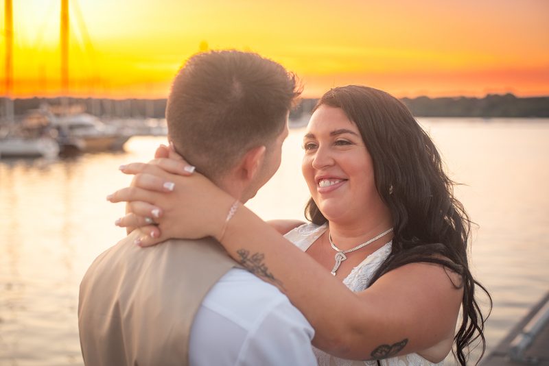 A couple embraces by the water at sunset, faces aglow with love and reflection from the golden light.